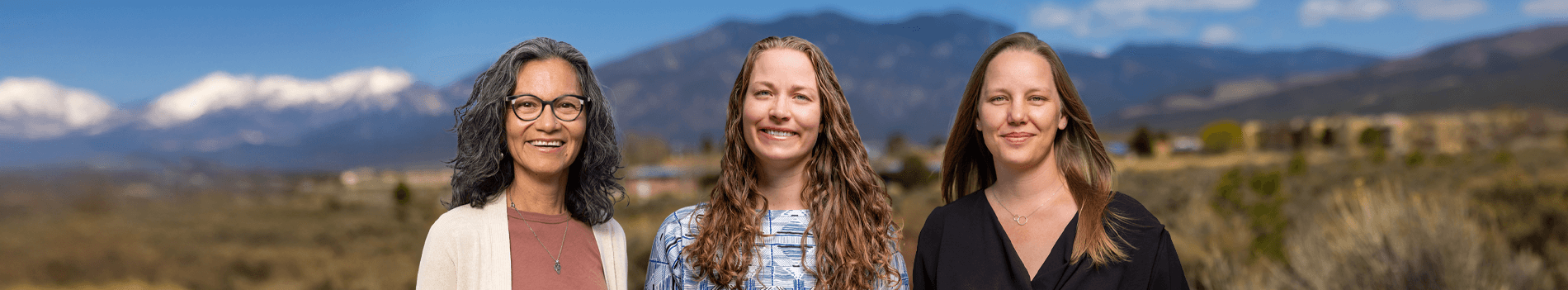 Three women stand together outdoors with mountains in the background, representing the Holy Cross Women’s Health Institute.