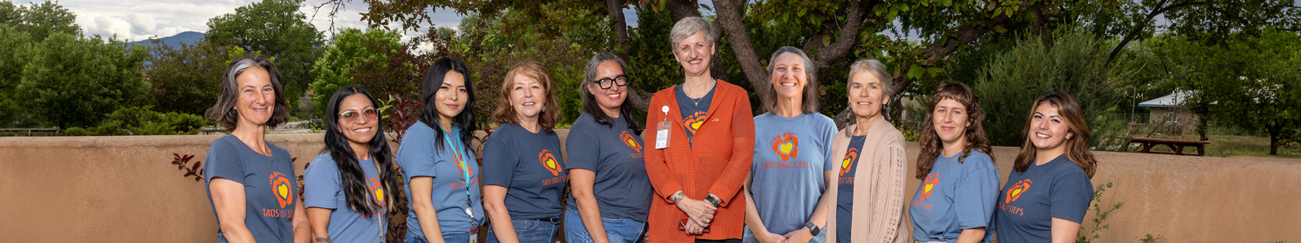 A group of diverse women in matching t-shirts stands together outdoors, smiling for a community-focused initiative.