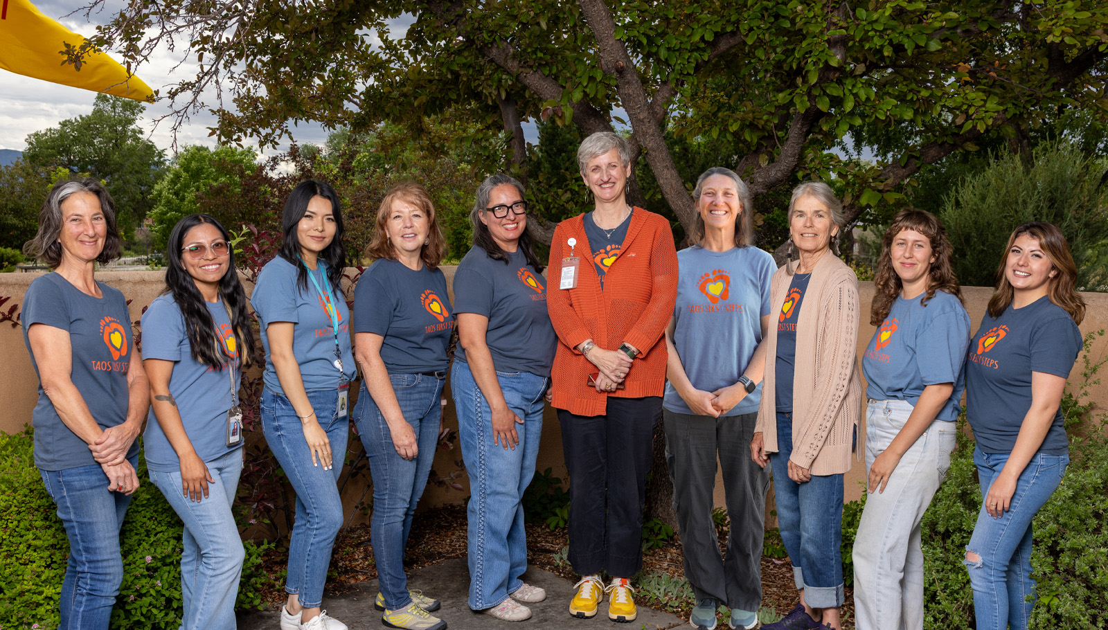 A diverse group of women in matching shirts poses outdoors, representing the "First Steps" initiative.