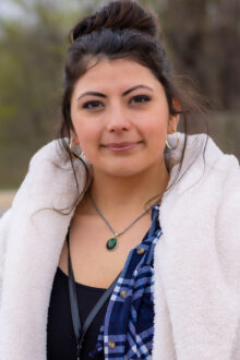 A woman with dark hair, wearing a cozy white jacket and plaid shirt, poses confidently outdoors.