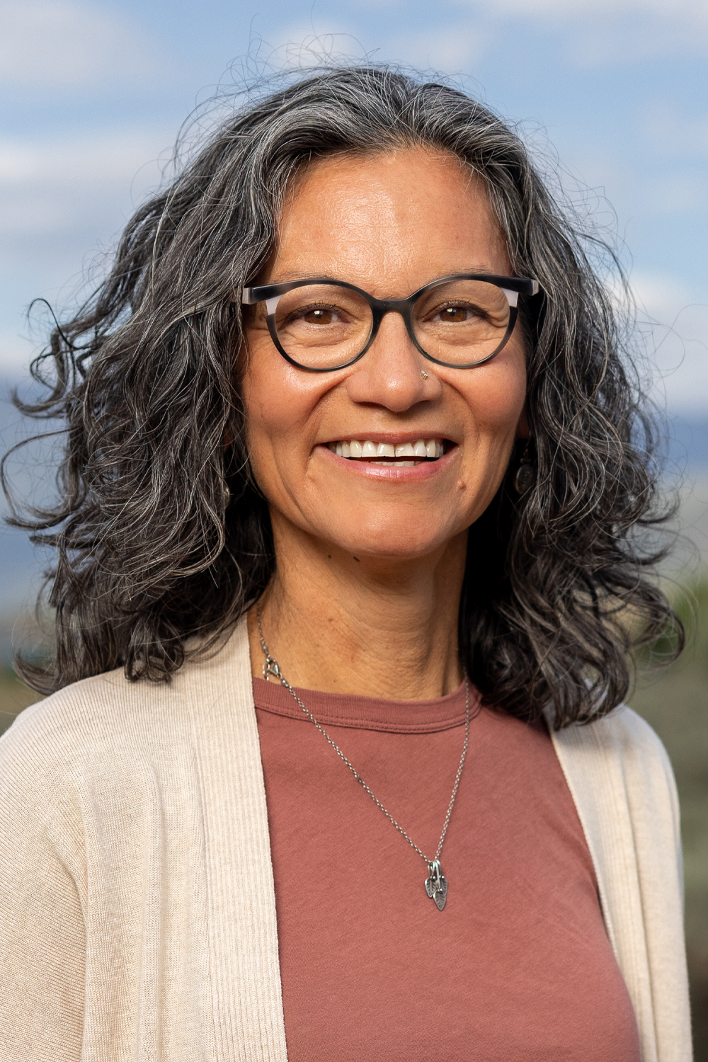 Smiling woman with curly gray hair, wearing glasses and a necklace, stands outdoors against a scenic background.