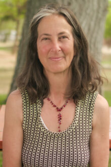 Beth Enson smiles warmly, wearing a patterned top and a necklace, against a natural outdoor backdrop.