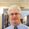 Michael Darnell, a middle-aged man with gray hair, wearing glasses and a blue dress shirt with a patterned tie, smiles warmly.