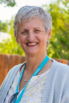 Smiling woman with short gray hair, wearing a light sweater and a blue lanyard, stands outdoors against a blurred background.
