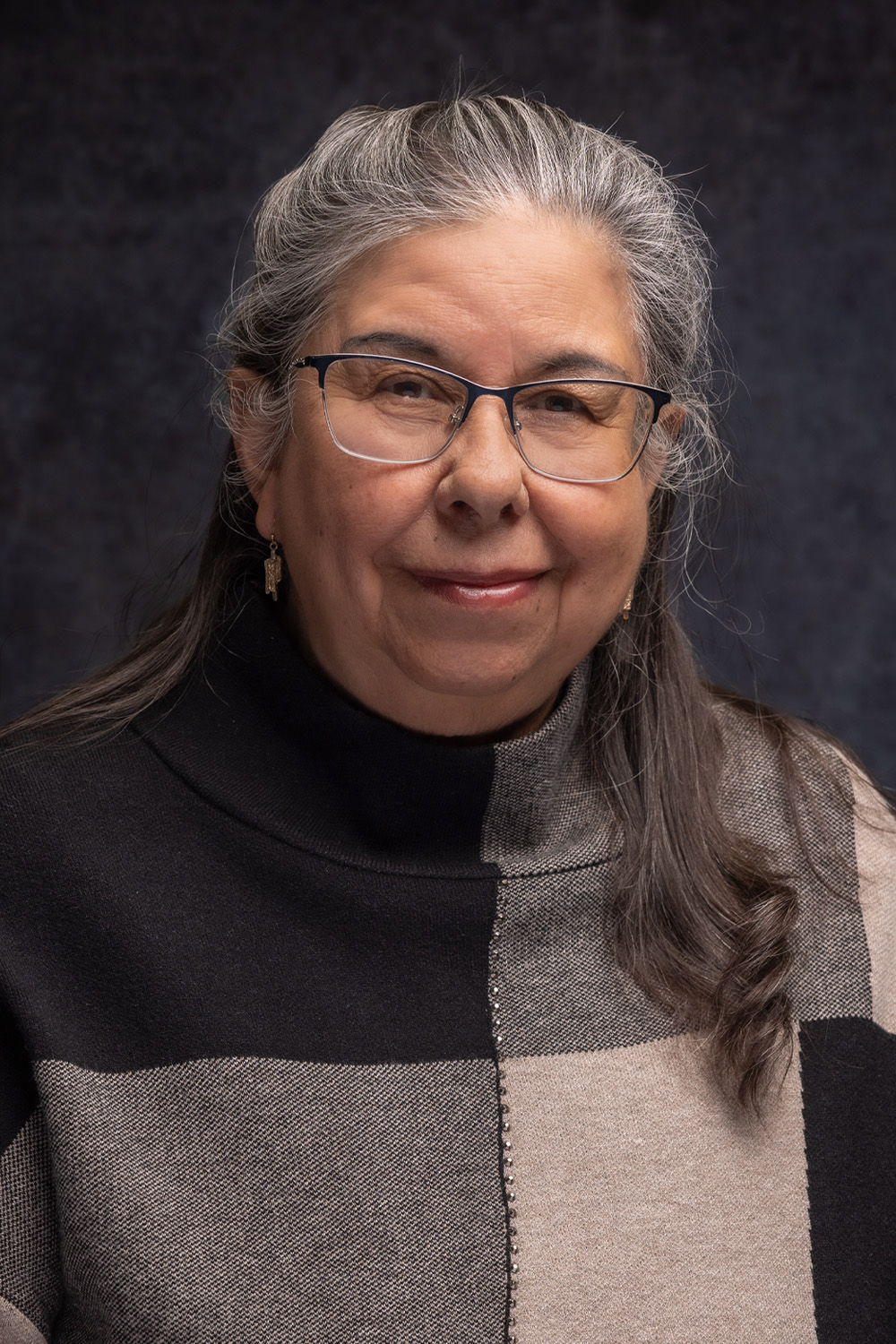 Lucille Gallegos-Jaramillo smiles warmly, wearing a stylish patterned sweater against a dark backdrop.