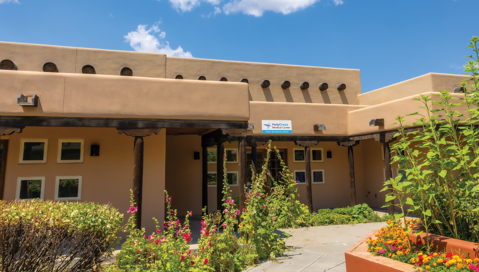 Holy Cross Primary Care features a welcoming adobe-style building surrounded by colorful flowers and greenery.