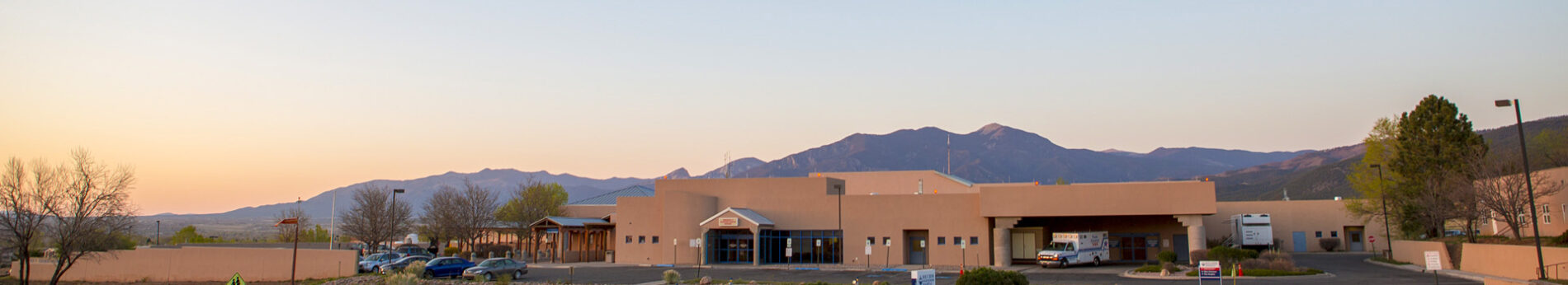 Laboratory building surrounded by mountains and trees, with vehicles parked in front during sunset.