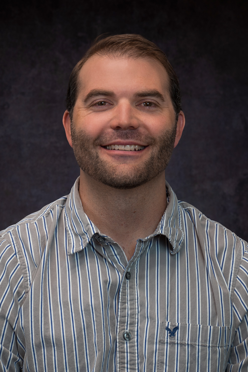 Smiling man with a beard wearing a striped shirt, set against a dark background, representing Jonathan Moran.