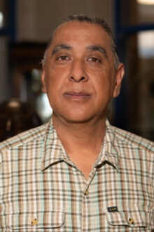 A middle-aged man with a neutral expression, wearing a plaid shirt, stands in a softly lit environment.