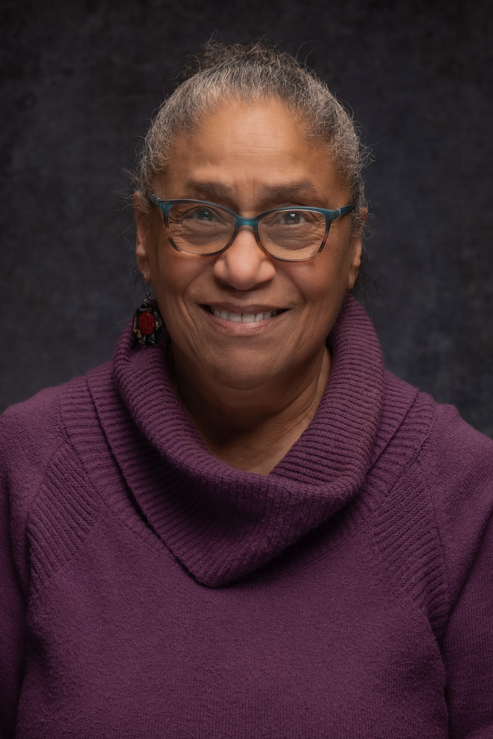 Smiling woman with glasses and earrings, wearing a purple sweater, against a dark background.