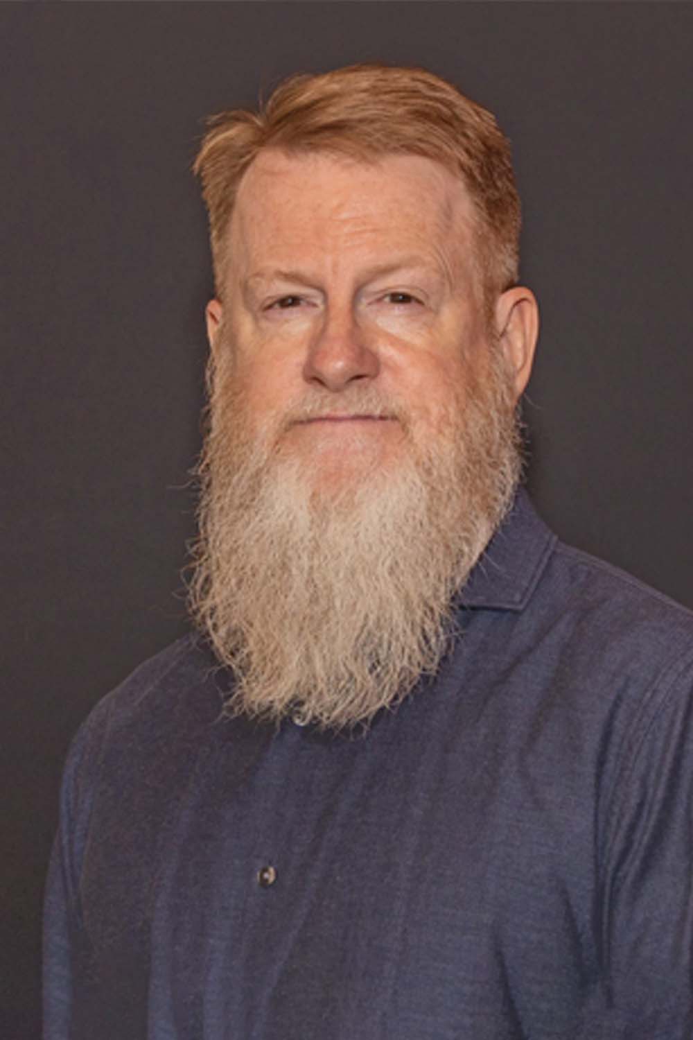Middle-aged man with a long beard, wearing a dark shirt, against a plain dark background.