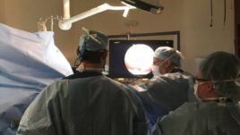 Surgeons in scrubs focus on a monitor displaying a surgical procedure in an operating room.