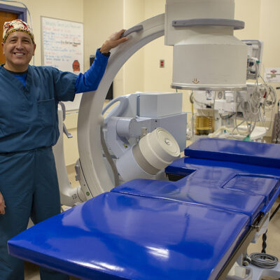 A smiling healthcare professional stands beside a modern medical imaging machine in a clinical setting.