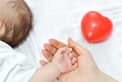 A baby's hand grasps an adult's hand beside a red heart-shaped toy, symbolizing care and connection in health initiatives.