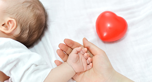 A baby's hand grasps an adult's hand beside a red heart-shaped toy, symbolizing care and connection in health initiatives.