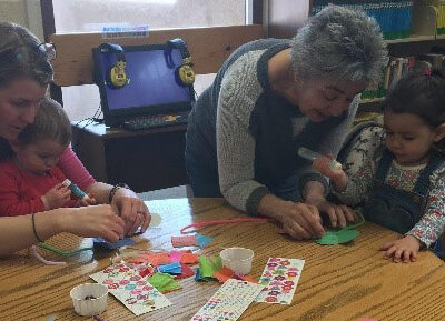 Adults and children engage in a crafting activity, creating art with colorful materials at a library table.