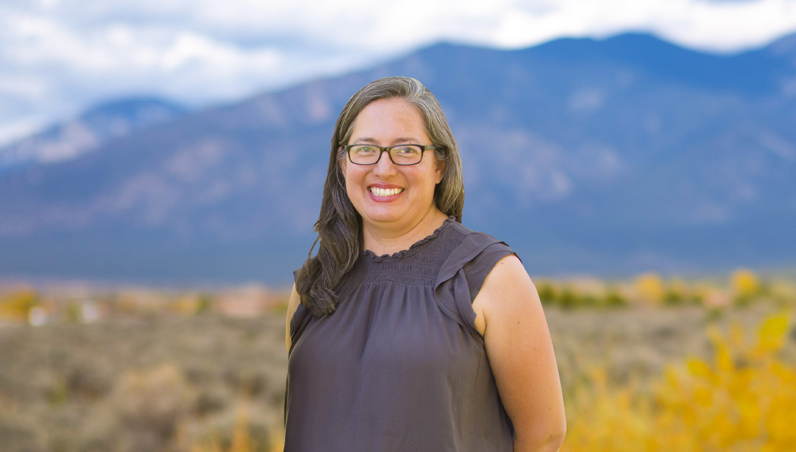 Smiling woman with glasses stands outdoors against a backdrop of mountains and autumn foliage, promoting community support for children.
