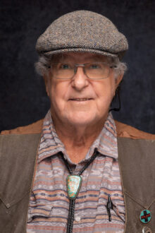 Elderly man with curly gray hair, wearing a patterned shirt, bolo tie, and cap, smiling against a dark background.