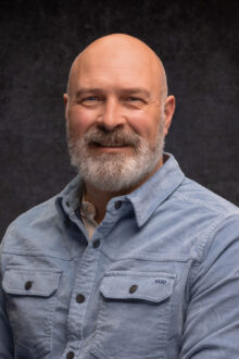 Smiling man with a beard wearing a light blue shirt, posing against a dark textured background.