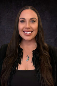 Robyn Chavez smiles confidently, wearing a black fringed jacket and a colorful pendant, against a dark background.