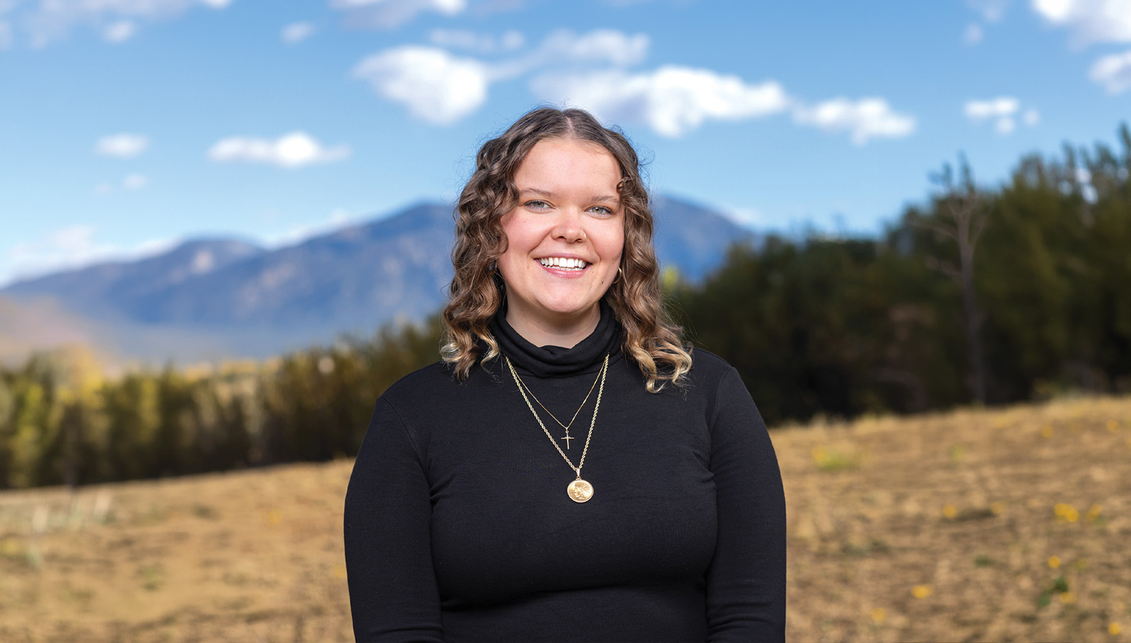 Smiling woman in a black turtleneck stands outdoors with mountains and trees in the background, promoting rural maternal services.