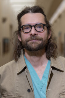 Dr. Adam Spring poses in a hallway, wearing a teal medical scrub and a beige jacket, with a friendly expression.