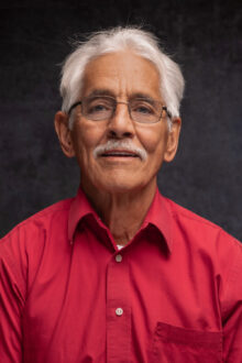 Elderly man with glasses and a mustache smiles warmly, wearing a red shirt against a dark background.