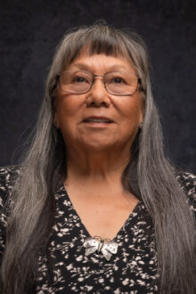Eileen Lujan, with long hair and glasses, wears a butterfly brooch and a patterned top against a dark background.