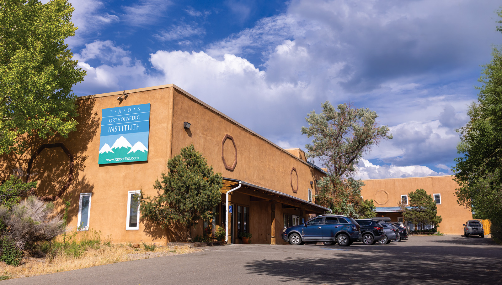 Taos Orthopedic Institute building with a sign, surrounded by trees and parked cars under a blue sky with clouds.