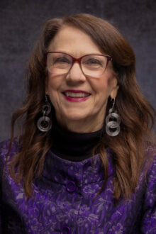 Smiling woman with long brown hair and glasses, wearing a purple patterned jacket and large silver earrings.