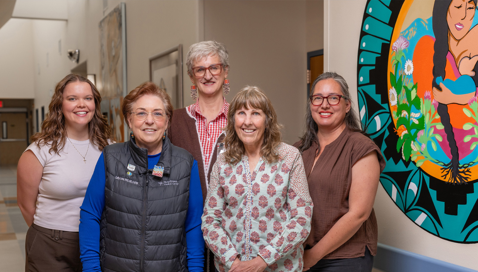 Five women stand together in a hallway, smiling, with a colorful mural depicting a nurturing figure in the background.