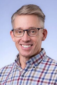 Smiling man with short hair and glasses, wearing a plaid shirt, against a light blue background.