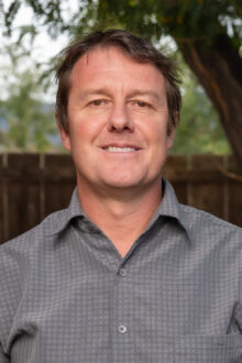 Portrait of Jason Pfeifer smiling outdoors, wearing a gray patterned shirt with a blurred natural background.