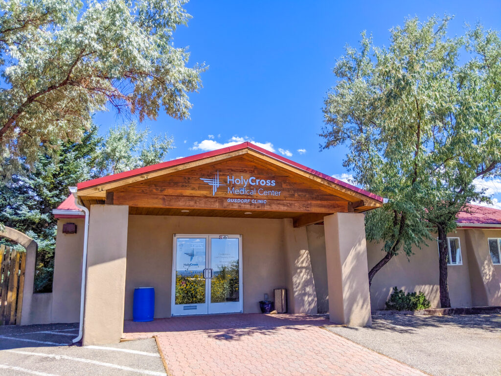 Holy Cross Medical Center's outdoor clinic entrance in Taos, surrounded by trees and a clear blue sky.