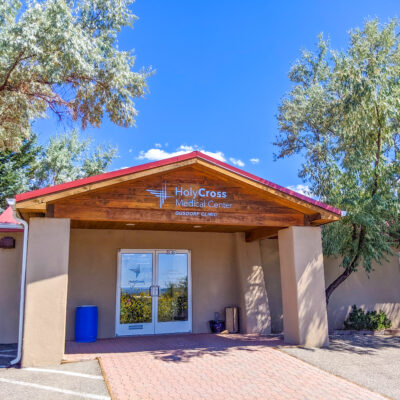 Holy Cross Medical Center's outdoor clinic entrance in Taos, surrounded by trees and a clear blue sky.