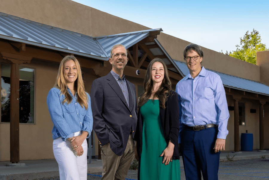 Four professionals stand together outside a building, smiling and dressed in business attire, representing a collaborative team.